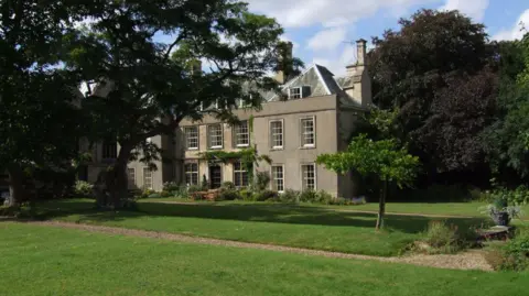 The National Trust The two storey house has large white windows and a third tier in the roof.  The second photo is taken from a slightly different angle and shows the trees in the gardens.