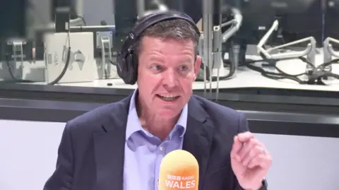 Rhun ap Iorwerth being interviewed in a Radio Wales studio, behind a microphone with an orange foam cover displaying the BBC Radio Wales logo. He is gesturing with his hand and wearing a pair of headphones over one ear. 