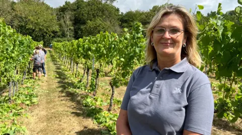 A woman with blonde hair stands in front of a series of grape vines.