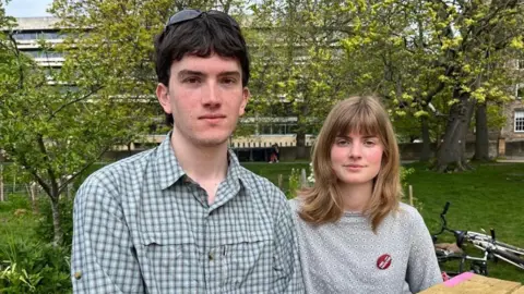 Rob, left, wearing a green checked shirt. He has dark hair and has sunglasses on his head. Fiona, right, wearing a grey jumper. She has shoulder-length brown hair. Both are staring straight at the camera.