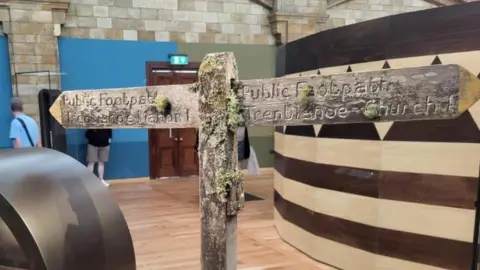 An image of an old-looking signpost in the Natural History Museum, featured with 'Public Footpath - Trentishoe Church' in one direction and 'Teddyshow Manor' in the other, adorned with moss or assumed lichens living on it. 