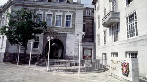 An exterior view of County Hall in Maidstone, a white Portland stone building with its name and the county crest on a slab in front of it, and four lampposts in front of a glass and metal set of doors.