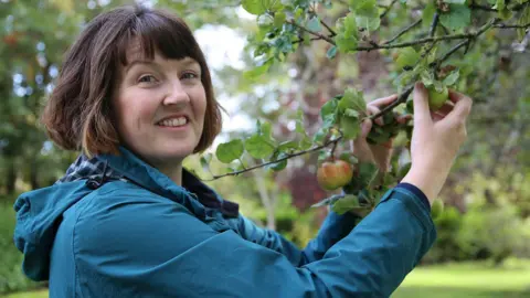 University of Worcester A woman with short dark hair, wearing a blue jacket, holds an apple tree branch and smiles at the camera. She is pictured side-on, from the shoulders up, with an orchard in the background, blurred.