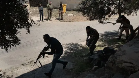 Getty Images Three men in military camouflage outfits carrying gun walk one after the other under some trees as several people watch on from the other side of the street