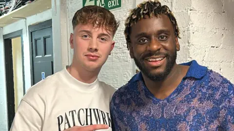 Kerr Mercer and Myles Smith smile for the camera while looking into it - Kerr has short brown hair and a white T-shirt on. Myles has a blue and oink patterned jumper.