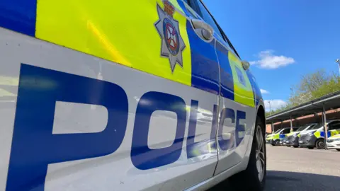 A close up shot of a Wiltshire Police car, showing the side, which says POLICE in large letters and has the force's symbol above it. There are other parked police vehicles in the background