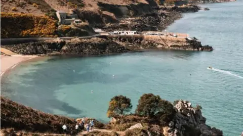 Jersey's Greve De Lecq beach, people are strolling down a nearby path and others are swimming in the sea. 
