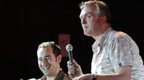 Stuart Steele/Civic Comedy A man in a short sleeved shirt holds a microphone upright in one hand. He is looking forwards, while a second man, sat lower than him, is looking at a piece of paper and smiling
