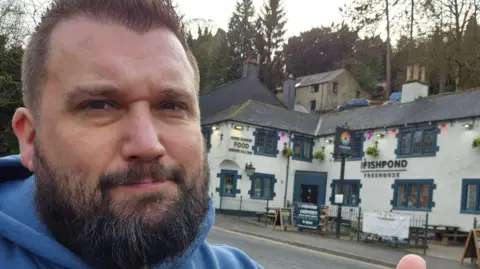 Dale takes a selfie outside a pub called The Fishpond. It's a white building with navy blue window frames 