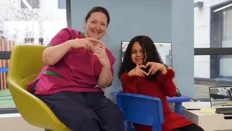 Senior health play therapist Charlotte Cooper sits in a yellow chair and is wearing a pink tunic. Rama, a 10-year-old girl, sits in a small blue chair on the right. She is wearing a red dress. Behind them is a computer screen showing Minecraft and a window, through which an outdoor play space is visible.