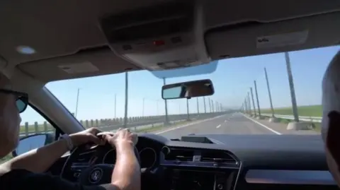 Lewis Inman Two people are sitting in the front of a moving vehicle. Ahead of them - through the windscreen, the road is long and straight and lives with large, high poles. The poles carry a large net acros the road, several feet above.
