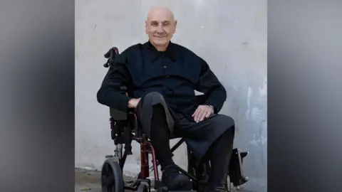 A smiling Bill Ward in black shirt, trousers and shoes, sits in a wheelchair against a plain grey background