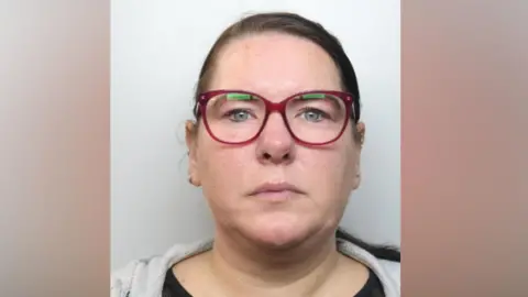 Thames Valley Police A police mug shot of a woman wearing glasses, a brown top and a grey jumper. She has brown hair which looks like it has been tied back in a pony tail.