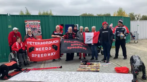 Shariqua Ahmed / BBC A group of people holding red Peterborough Speedway flags with a panther statue on the ground in front of them. 