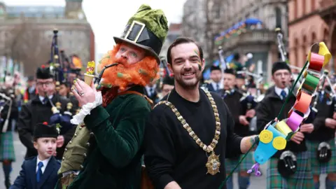 Lord Mayor of Belfast Micky Murray, wearing a black jumper and his gold chain of office, poses for a photo at the start of Belfast's St Patrick's Day parade.   He is standing back-to-back with a man in a green velvet leprechaun suit, large green hat and a fake ginger beard.   He is pretending to smoke a packet of cheese and onion crisps from a pipe. Bagpipe players are marching behind them. 