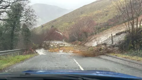 Landslide seen coming onto the A487 road between Corris and Minfford. A heavy flow of water can be seen flowing down the mountain across the road, with trees and debris. 