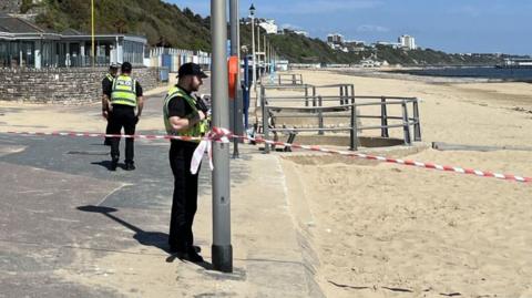 Police officer stands by sandy beach and cordon