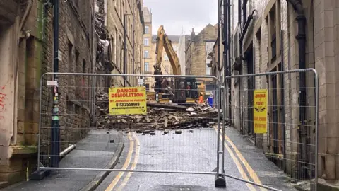 A yellow mechanical digger behind metal fencing lifting debris.