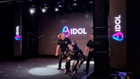 Two men dressed in black rush towards a grey robot toppling forward.on stage as it topp