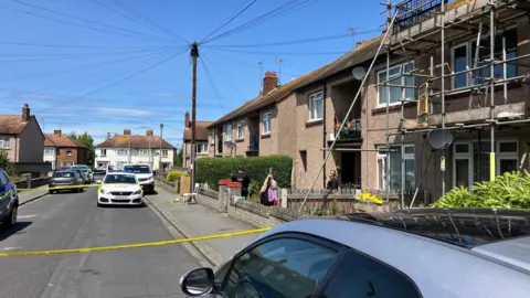 An image of the residential street where the incident happened. Yellow police tape has blocked off the house involved. A police car can be seen behind the tape and two officers are standing in front of the property. 
