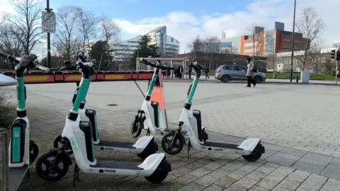 Several rental e‑scooters are parked on a paved area near a road, with modern hospital buildings and trees in the background.
