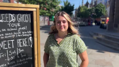 A picture of Daisy Lyons, who has blonde hair and is wearing a green and white top, standing nest to her ghost tours sign, there are buildings and trees in the background.