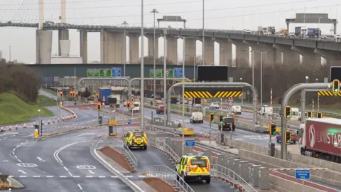 An image of traffic waiting to join one of the bore tunnels at the Dartford Crossing. In the background, there is the QEII Bridge. 