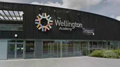 Google maps street view of Wellington Academy. The building is wide and covered in black wooden panels, with a large Wellington Academy logo on the front.