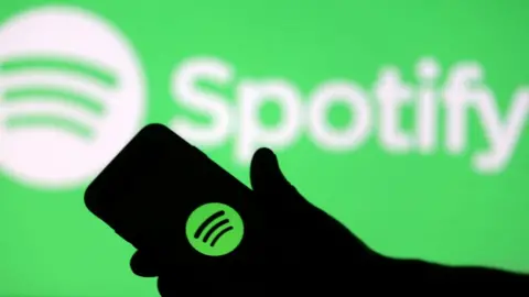 रॉयटर्स एक स्मार्टफोन को Spotify लोगो के स्क्रीन प्रक्षेपण के सामने देखा जाता है