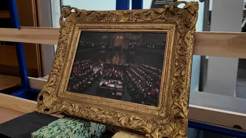 A painting of politicians taking part in a debate. The painting appears in a gold frame 