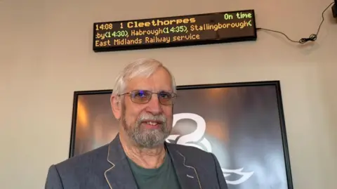 The photo shows a man smiling at the camera. He's wearing a blue blazer, a green top and glasses. He's standing in front of a black frame. Above his head is a train departure board, which reads "Cleethorpes" and various other railway stations in LED lighting. 