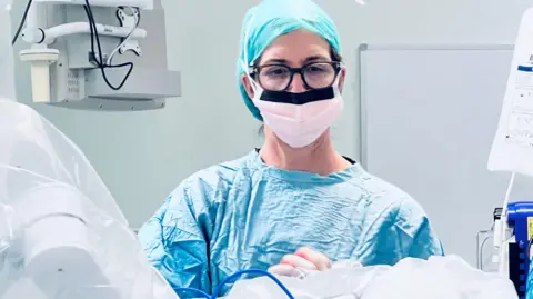 Caroline Burt stands in an operating theatre. She is wearing blue scrubs and a white face mask, and glasses.