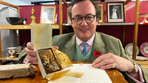 John Fairhall/BBC Auctioneer Jonathan Benson holding the cake, which looks mouldy. There is a letter in front of him and he is sat in front of various portraits of Queen Elizabeth II and Prince Philip, as well as china. Mr Benson is wearing a suit with a jumper, pink shirt and tie underneath and a poppy.