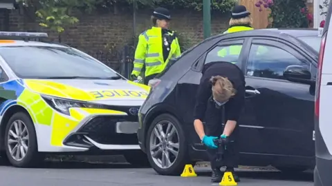 A forensics officer taking photos of a crime scene. There is a black car and a police car behind the officer, as well as two police officers.