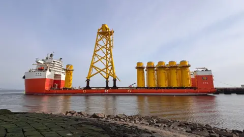 The heavy lift vessel Hua Yang Long sitting in port at Leith. It is mainly red and white but has a large yellow tower in the middle as well as large yellow cylinders.