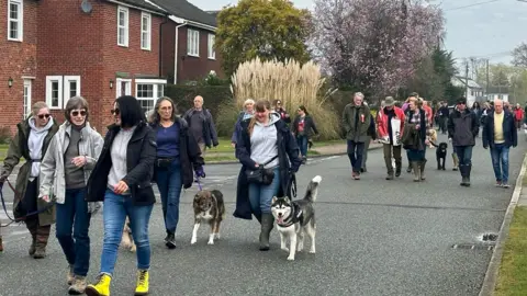 Jenny Kirk/BBC About 20 people walking along a residential road, plus two dogs, with housing behind them 