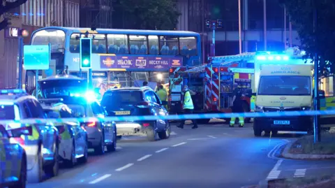 A night-time shot showing a bus, ambulance, fire engine and several police cars and police tapes on a town centre street 