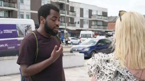 A man wearing a maroon t shirt wearing a backpack stands on a street corner gesturing with his right hand while talking to a blonde woman, in front of a busy road with shops behind 