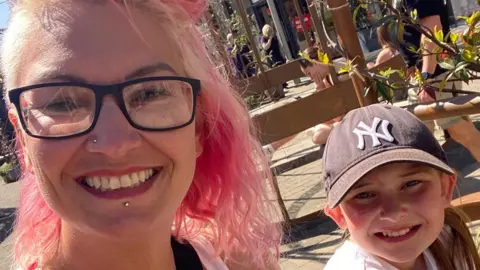 Charlotte Charlotte with pink hair and dark rimmed glasses with her daughter Bella who is wearing a baseball cap. They are both smiling at the camera.