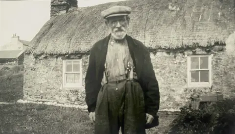 Manx National Heritage) Black and white photo of an older man standing in front of a thatched cottage.