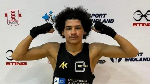 Top Yard School of Boxing Ismael wearing a black vest and black shorts and posing with his hands curled up next to him