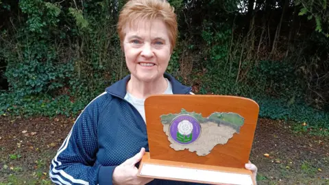 Hilary King A woman with short red hair smiles as she holds up an old fashioned wooden tropy. She's wearing a navy tracksuit jacket.
