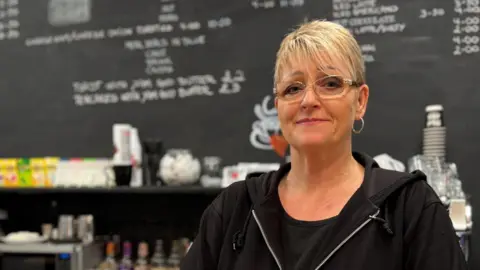 Sharon Edwards/BBC Anne Britt stands at the counter of her cafe. She has short blonde hair and wears glasses, gold-ring earrings, a black T-shirt and a black hoodie. Behind her, the menu is written in white chalk on a large blackboard.