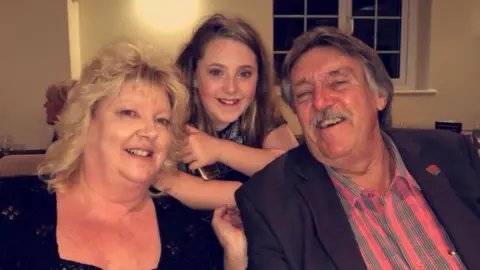 Vicky Richards Ashleigh has long dark hair and is standing behind Lorraine and John, who are seated. Lorraine is wearing a black, square-neck top and has shoulder length blonde hair. John is wearing a red and grey striped shirt and a black blazer. He has grey hair.