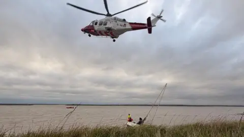 A red and white helicopter flying in the sky, over water, with two people in a white boat that is listing. Another red vessel is in the water, and there are reeds in front of the water. A cable is going from the helicopter to the boat. 