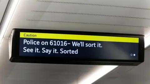 An electronic display inside a London Underground train showing a yellow “Caution” banner and a message advising passengers to contact police on 61016, alongside the slogan “See it. Say it. Sorted”.
