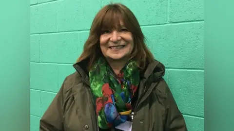 LDRS Jill Hood has shoulder-length brown hair with a fringe. She is standing in front of a green wall. She is wearing a khaki green coat with a green scarf that has red, blue and yellow flowers on it.