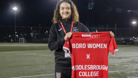 Sammie Leigh is dressed in black football kit and she holds footballs and a t-shirt reading &#x27;BORO WOMEN X MIDDLESBROUGH COLLEGE&#x27;. She is standing on a football pitch at night. 