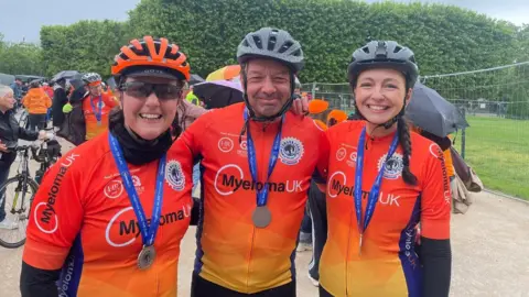 Deb Gascoyne The woman from the above picture, wearing the same lycra as before. She is wearing sunglasses and an orange cycle helmet and has a medal hanging around her neck on a blue ribbon. On her left are a middle-aged man and a young woman, all wearing the same lycra and with the same medals.