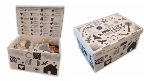 Rotherham Council On the left is an open black-and-white patterned baby box. Items inside include a cuddly grey rabbit with yellow ears, and white and grey socks. Under the lid is a diagram of the box's contents. On the right is a closed box with pictures on top of nature and wildlife.